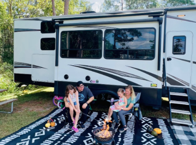 Family sitting around a campfire outside a travel trailer with slide-out extended at a wooded campsite.