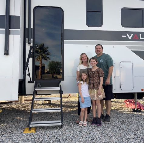 Family of four standing beside a travel trailer RV near the entry steps at a campground.