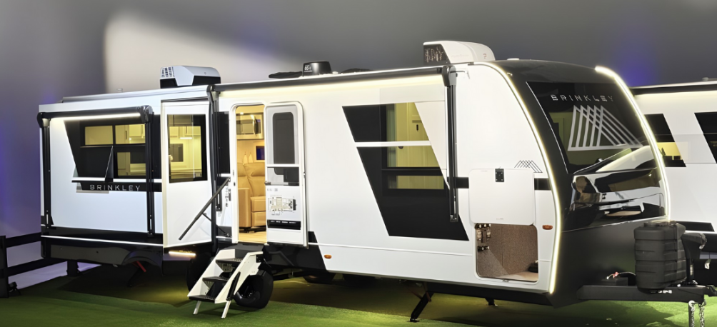 Brinkley travel trailer RV with slide-out extended and entry steps down, showcasing the modern exterior design and illuminated interior at an RV show.