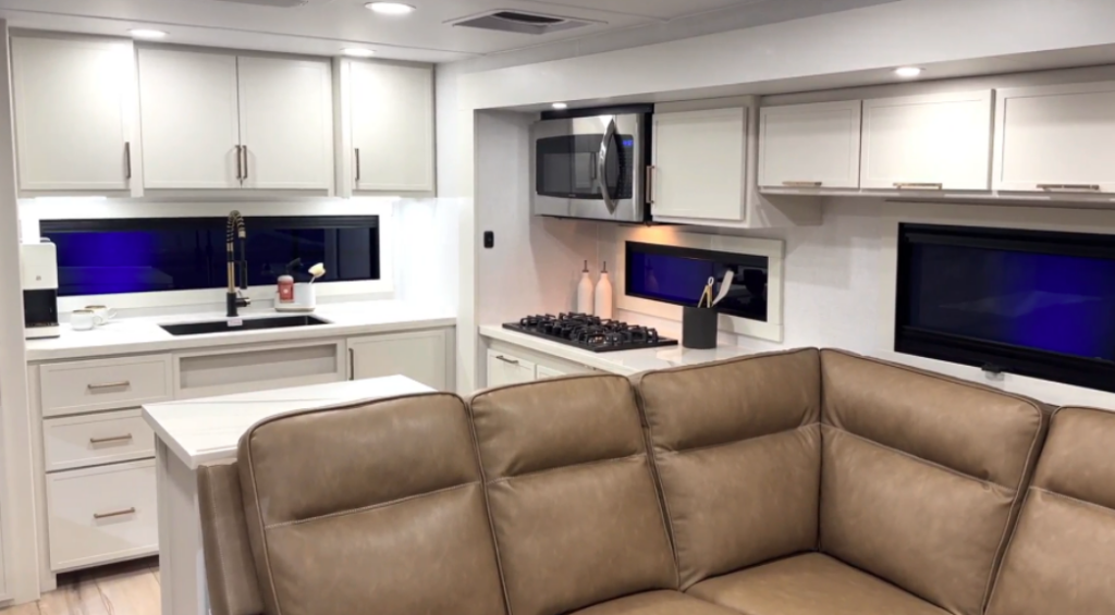Modern RV interior featuring a kitchen with white cabinetry, microwave, gas cooktop, sink, and an L-shaped sofa seating area.