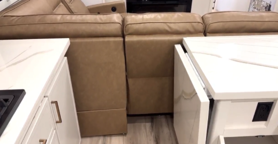RV kitchen island with pull-out countertop extension and storage beside a brown leather sofa in a modern travel trailer interior.