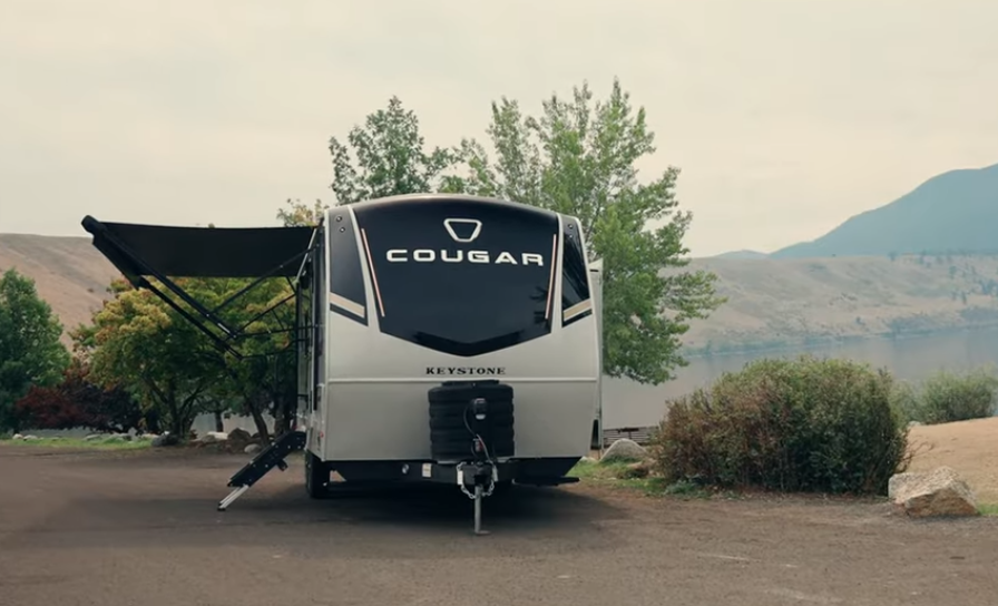 Keystone Cougar travel trailer parked at a scenic lakeside campsite with mountains in the background and awning extended.