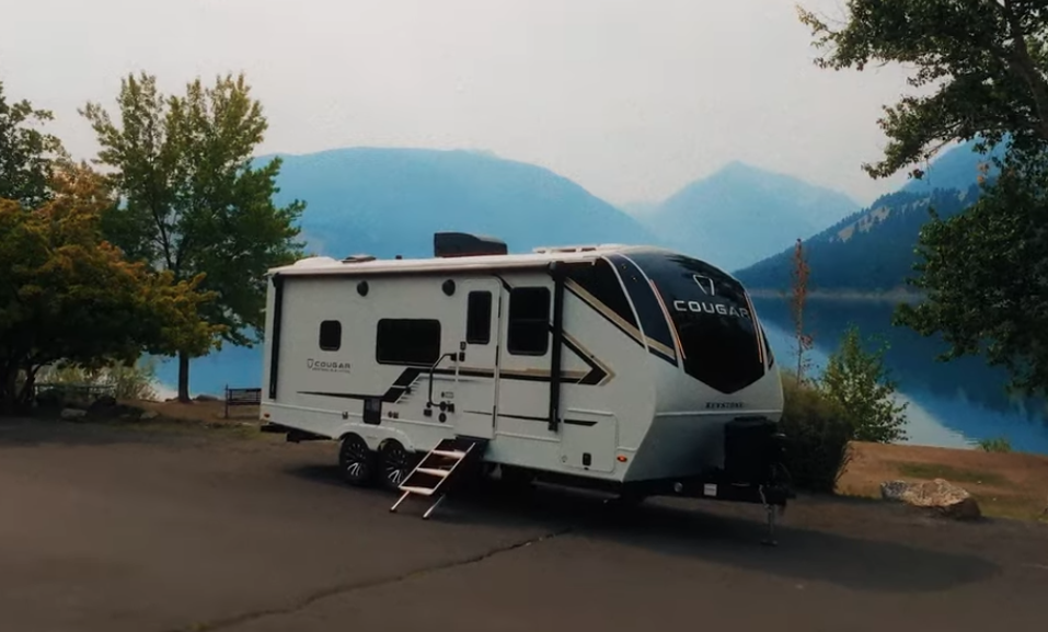 Keystone Cougar travel trailer parked at a lakeside campsite with mountain views and trees in the background.