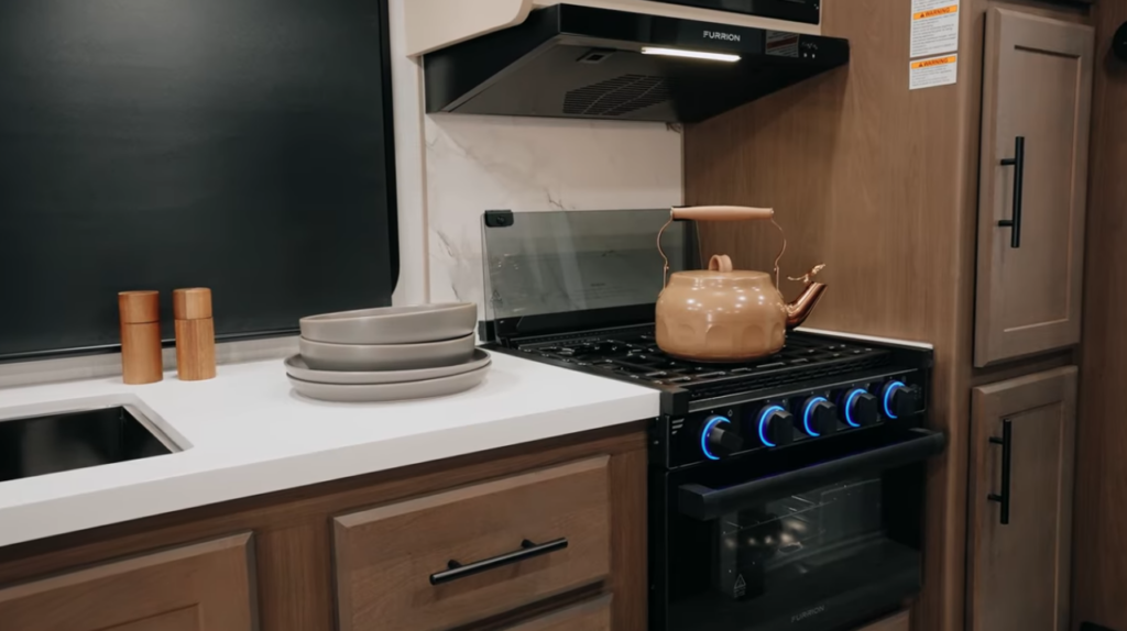 RV kitchen with gas range and oven, teakettle on stovetop, countertop with dishes, and wood cabinetry in a modern travel trailer interior.