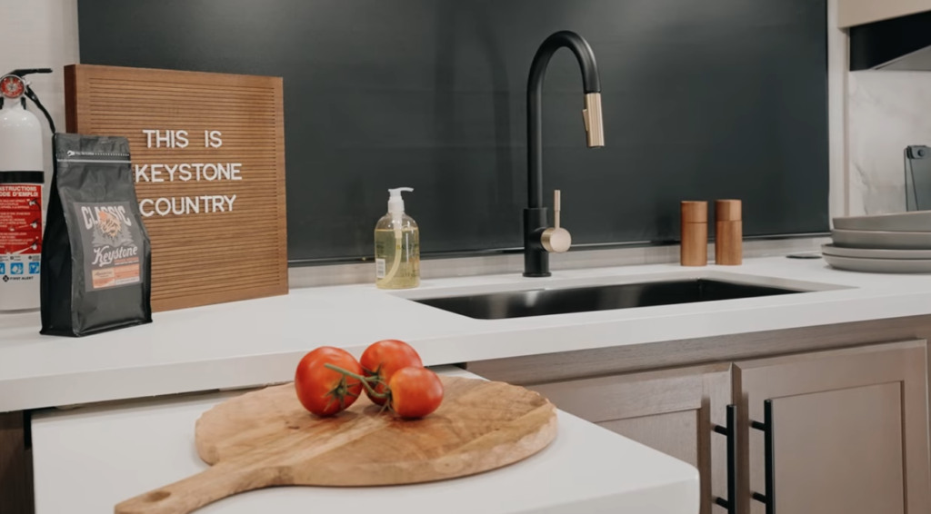 Modern RV kitchen sink with matte black faucet, countertop cutting board with tomatoes, and Keystone décor in a travel trailer interior.