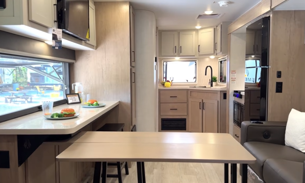 Modern travel trailer interior with kitchen, bar-style dining counter, freestanding table, and light wood cabinetry.