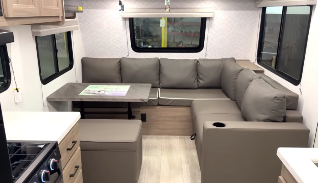 Travel trailer living area with U-shaped sofa seating, dinette table, and large windows inside a modern RV interior.