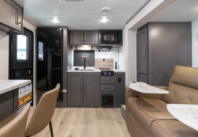 Compact travel trailer interior with gray kitchen cabinetry, microwave, refrigerator, and small dinette seating area.