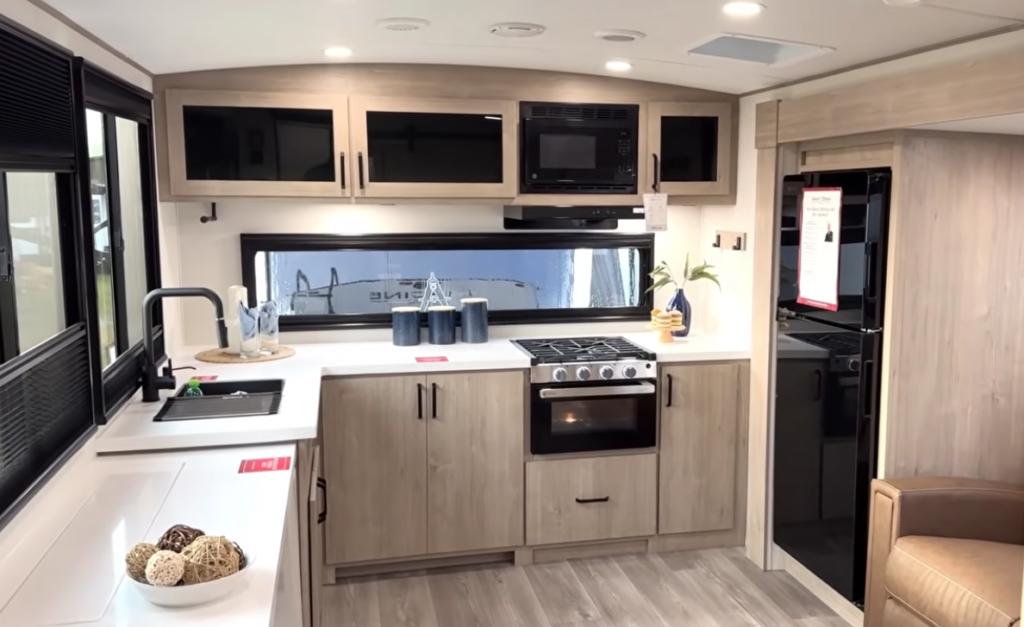 Modern travel trailer kitchen with gas range, microwave, large window backsplash, sink, and light wood cabinetry.