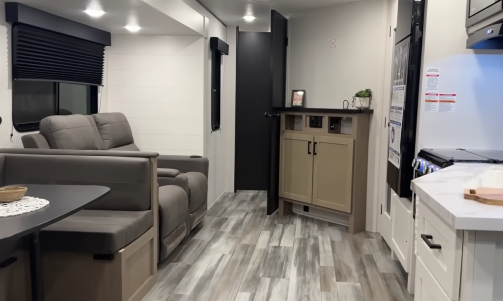 Travel trailer interior with theater seating, booth dinette, kitchen counter, and hallway leading to bathroom and bedroom.