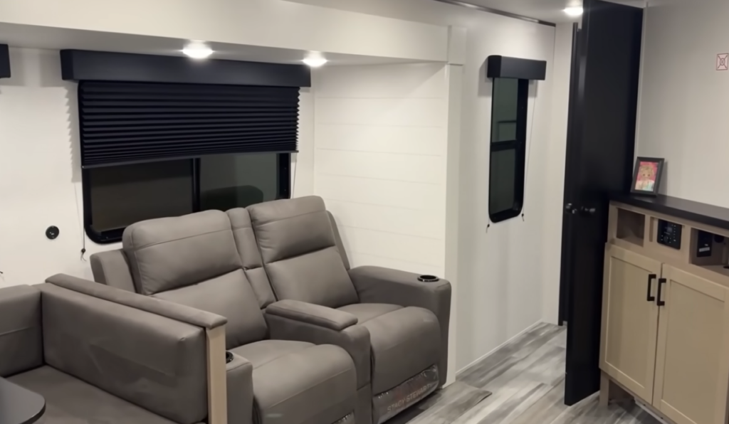 Travel trailer living area with dual reclining theater seats, window shades, and storage cabinet near hallway.