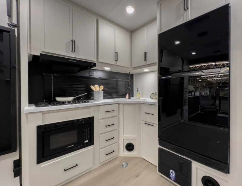 Travel trailer kitchen with white cabinetry, gas cooktop, microwave, large refrigerator, and corner countertop workspace.