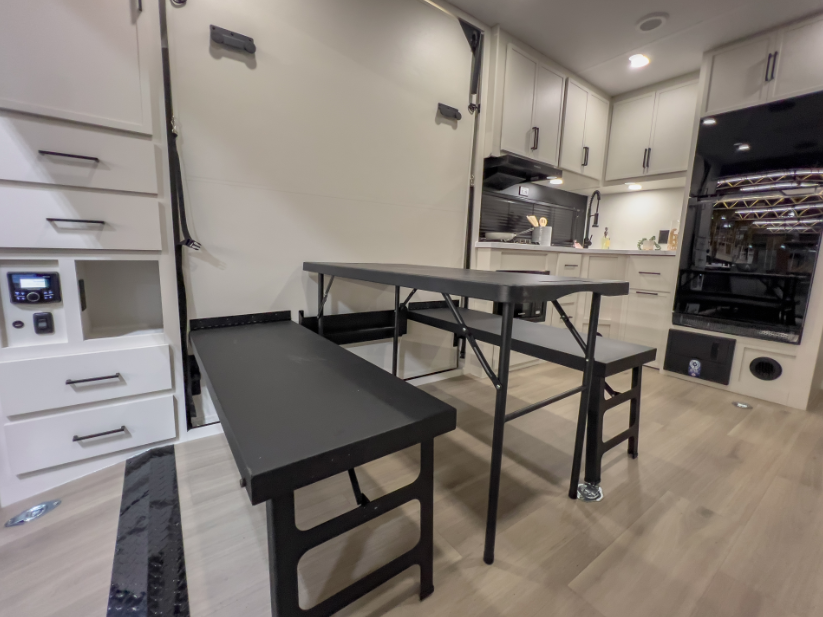 Toy hauler travel trailer interior with folding dinette table and benches, garage wall, and kitchen with white cabinetry.