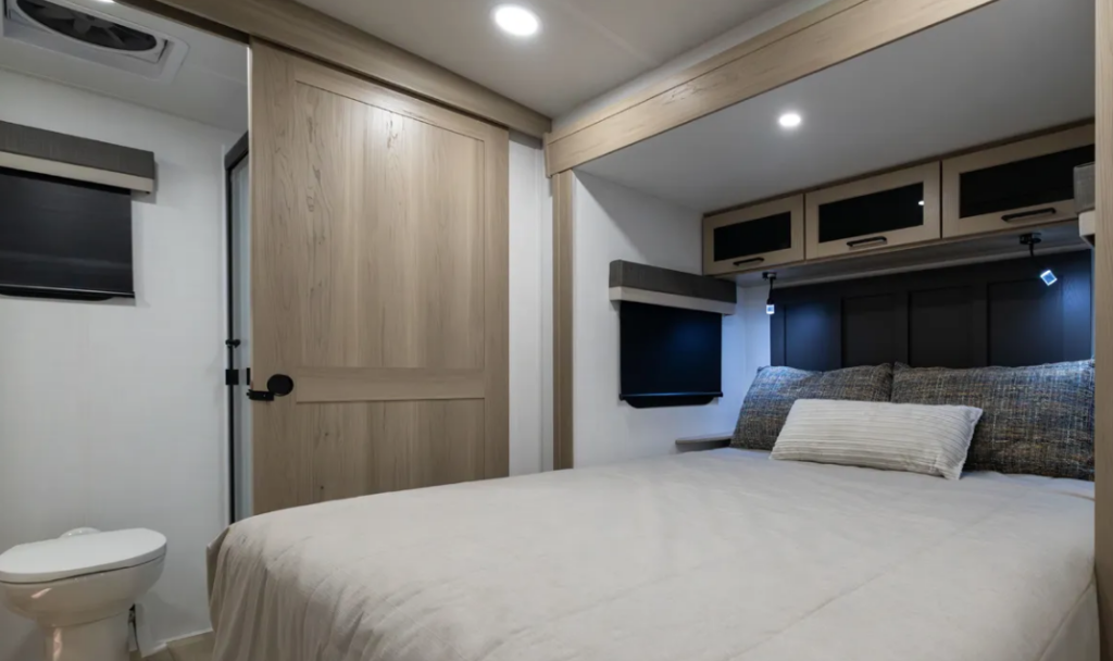 Travel trailer bedroom with queen bed, overhead storage cabinets, reading lights, and sliding barn door to bathroom.