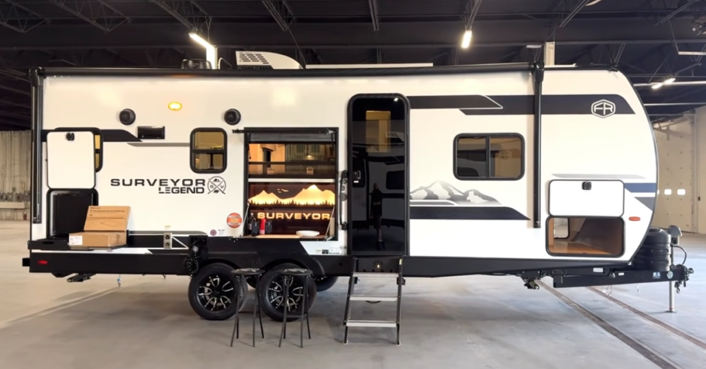 Forest River Surveyor Legend travel trailer with outdoor kitchen and entry steps displayed inside an RV warehouse.