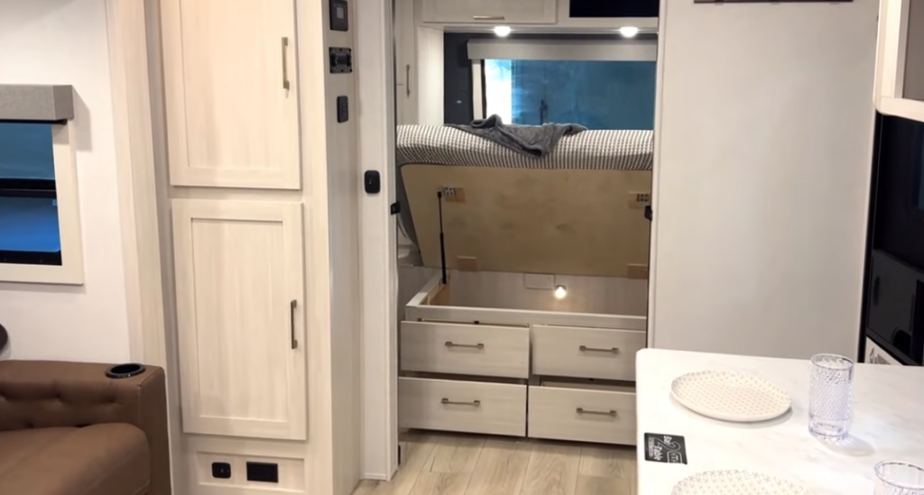 Travel trailer bedroom with lift-up bed storage and built-in drawers, showing organized under-bed storage space.