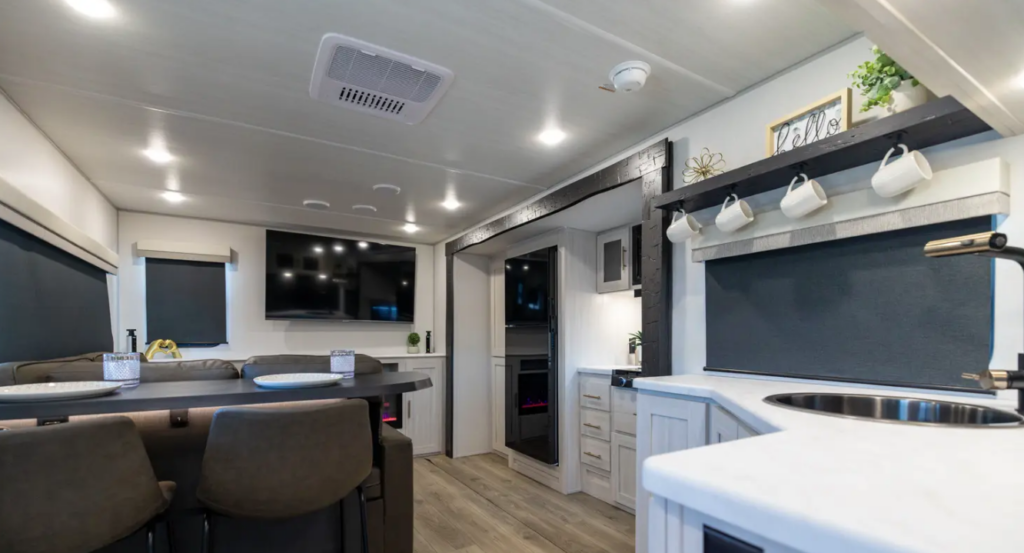 Travel trailer interior with bar-style dinette, kitchen sink and counter, wall-mounted TV, and modern farmhouse décor.