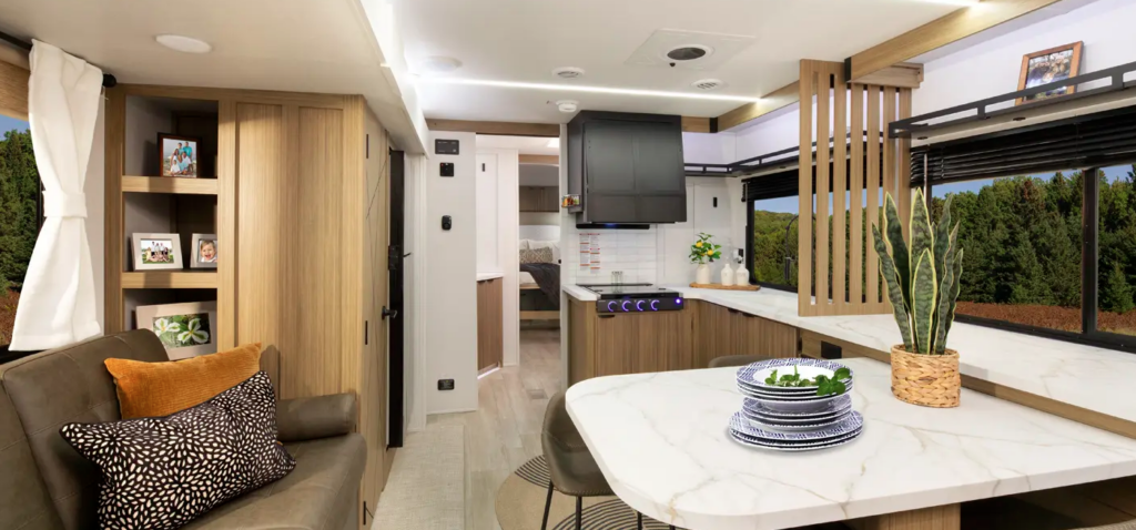 Contemporary travel trailer interior with kitchen, dining table, sofa seating, and hallway leading to the bedroom.