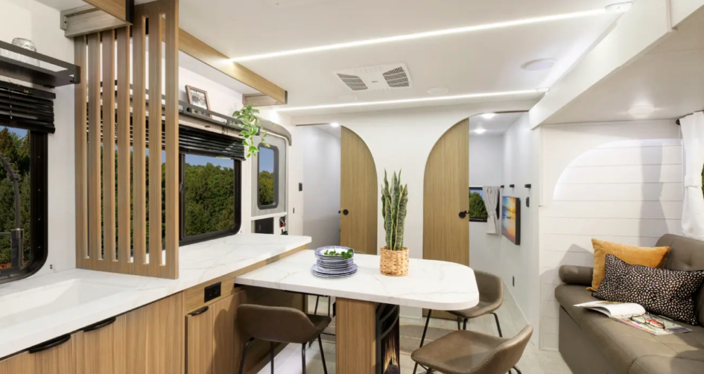Modern travel trailer interior with curved wood doors, dining table with chairs, sofa seating, and contemporary kitchen counter.
