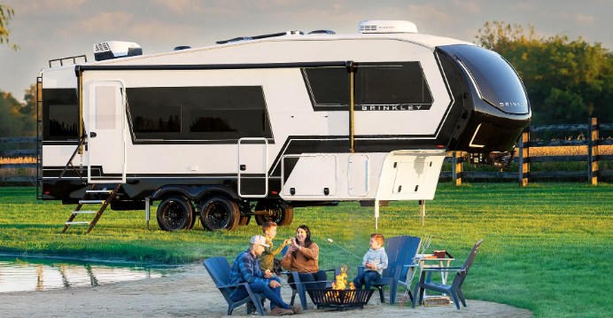 Family sitting around a campfire beside a Brinkley fifth wheel RV parked near a pond at sunset.