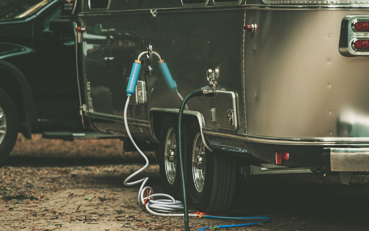 Travel trailer connected to water and power hookups at a campsite with hoses and cables attached near the wheel.