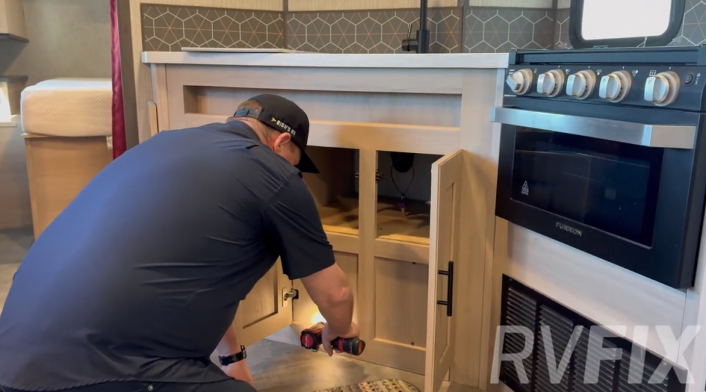 RV technician inspecting under-sink plumbing in a travel trailer kitchen with cabinet doors open and tools in hand.