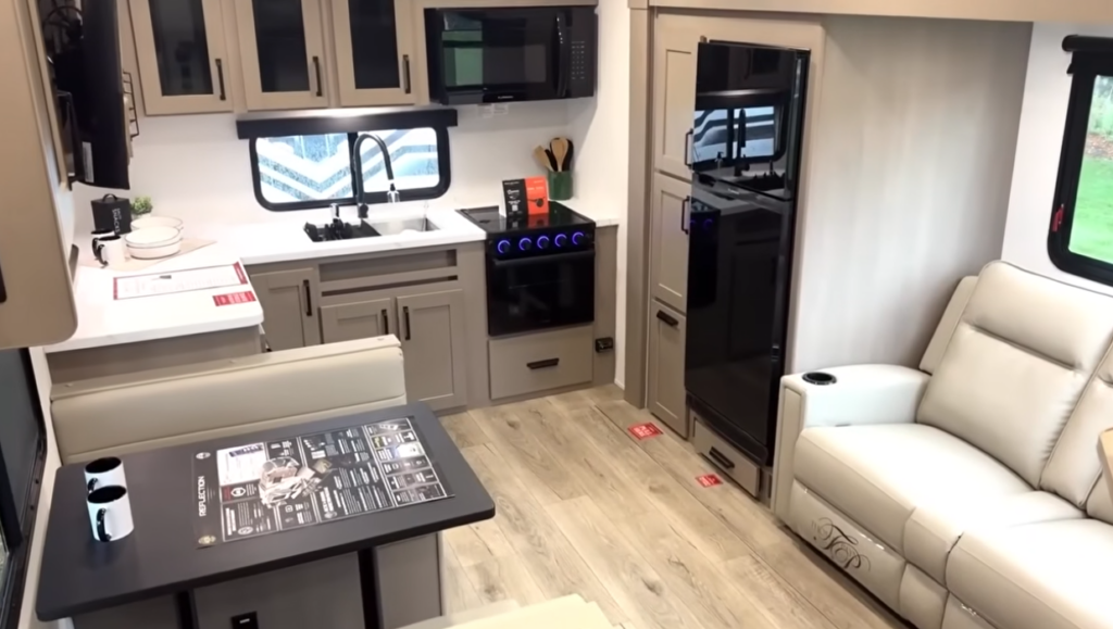 Compact travel trailer interior with kitchen, dinette table, sofa seating, and refrigerator in a modern RV layout.