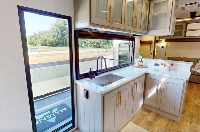 Travel trailer kitchen with farmhouse sink, large windows, white cabinetry, and natural light from campsite view outside.
