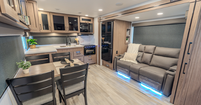 Modern travel trailer interior with a dinette table and chairs, full kitchen with wood cabinetry, and theater seating with blue accent lighting in a spacious slide-out living area.