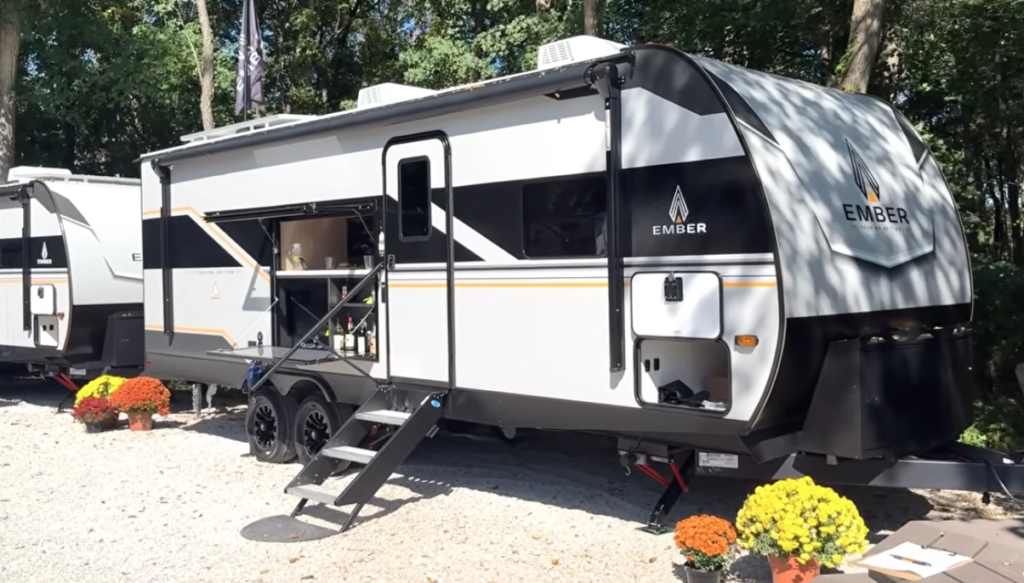 Ember travel trailer parked outdoors with slide-out extended, entry steps down, and an open exterior kitchen compartment, surrounded by trees and decorative flowers.