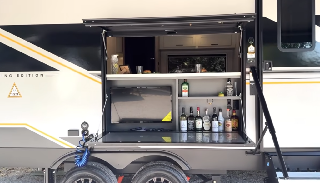 Travel trailer exterior with an open outdoor kitchen compartment featuring a TV, mini fridge, shelves with bottles, and a fold-up serving counter above dual axles.