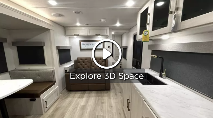 Modern travel trailer interior with a dinette booth, recliner seating, and a sleek kitchen featuring white countertops, overhead cabinets, and a large sink, shown with a 3D tour overlay.