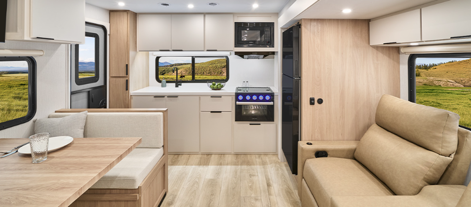 Bright, modern travel trailer interior with a dinette, sofa seating, and full kitchen featuring light wood cabinetry and large windows with scenic outdoor views.