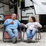 Couple relaxing in reclining camp chairs outside their travel trailer, holding hands and enjoying drinks at a campsite.