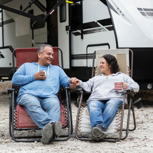 Couple relaxing in reclining camp chairs outside their travel trailer, holding hands and enjoying drinks at a campsite.