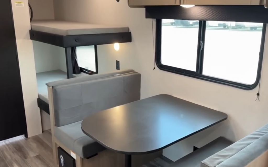 Compact travel trailer interior with a booth dinette next to a window and rear bunk beds with built-in lighting.