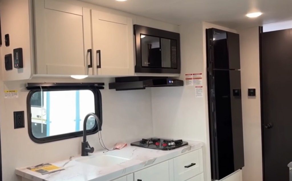 Travel trailer kitchen with white cabinetry, microwave, stovetop, black refrigerator, and a sink beneath a window.