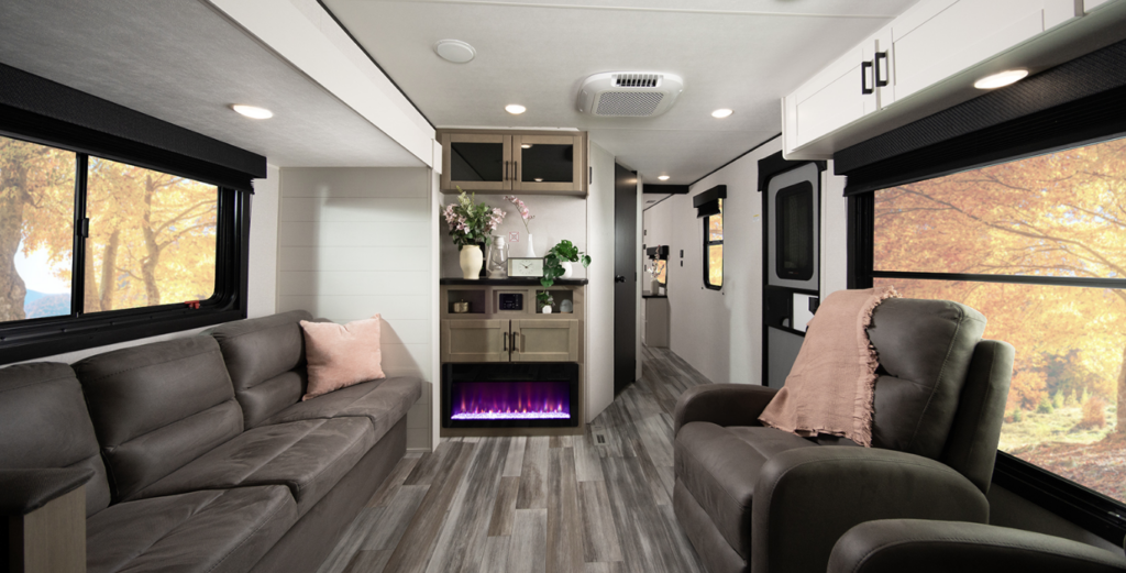 Travel trailer living area with a sofa and recliner, electric fireplace, storage cabinet, and large windows showing autumn scenery outside.