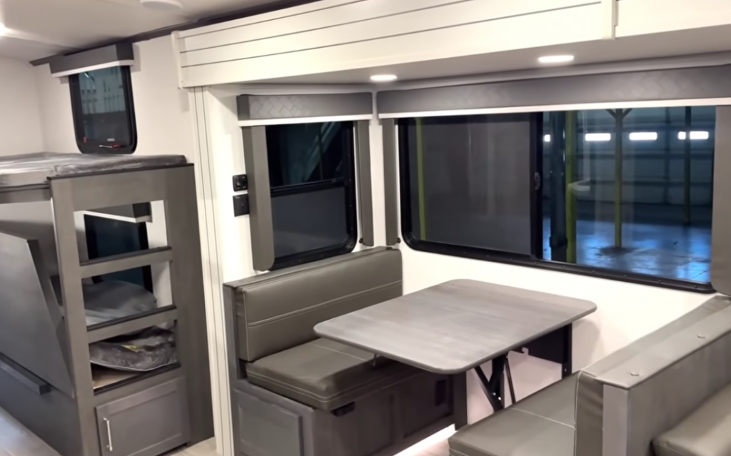 Travel trailer dinette with bench seating and a rectangular table, positioned beside large windows, with bunk beds visible in the background.