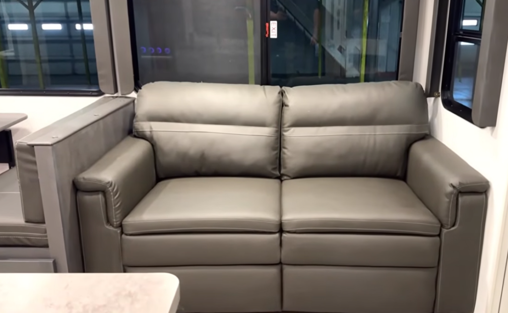 Compact loveseat sofa inside a travel trailer, positioned against large windows with surrounding cabinetry and seating.