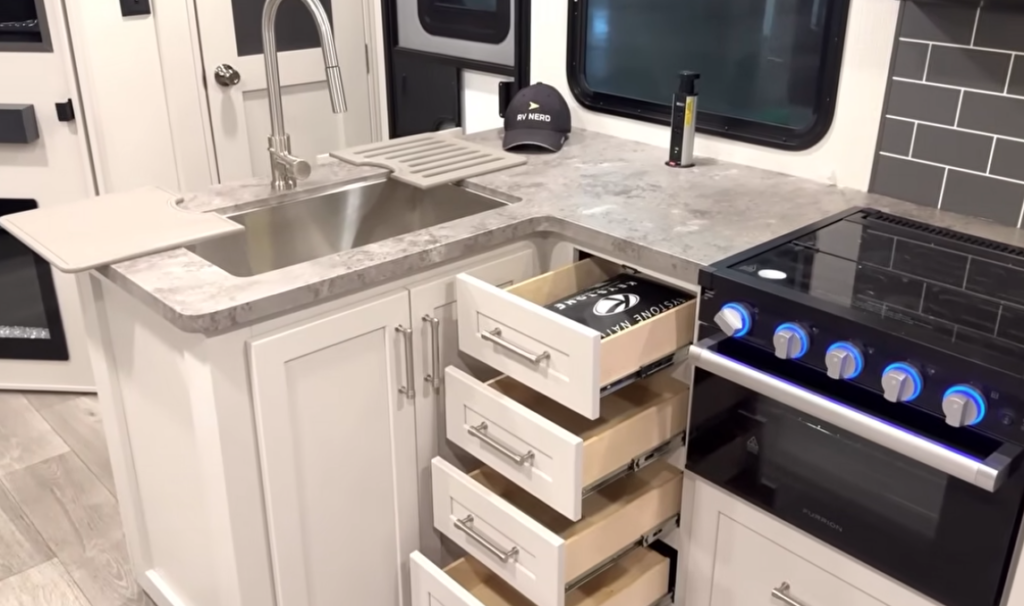 Travel trailer kitchen with corner sink, solid-surface countertops, stove with blue-lit controls, and multiple pull-out drawers for storage.
