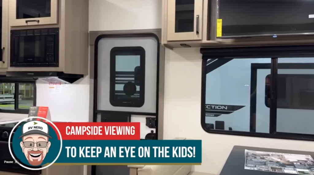 Travel trailer interior showing kitchen cabinets and a window with campsite visibility, with text overlay reading “Campside viewing to keep an eye on the kids!