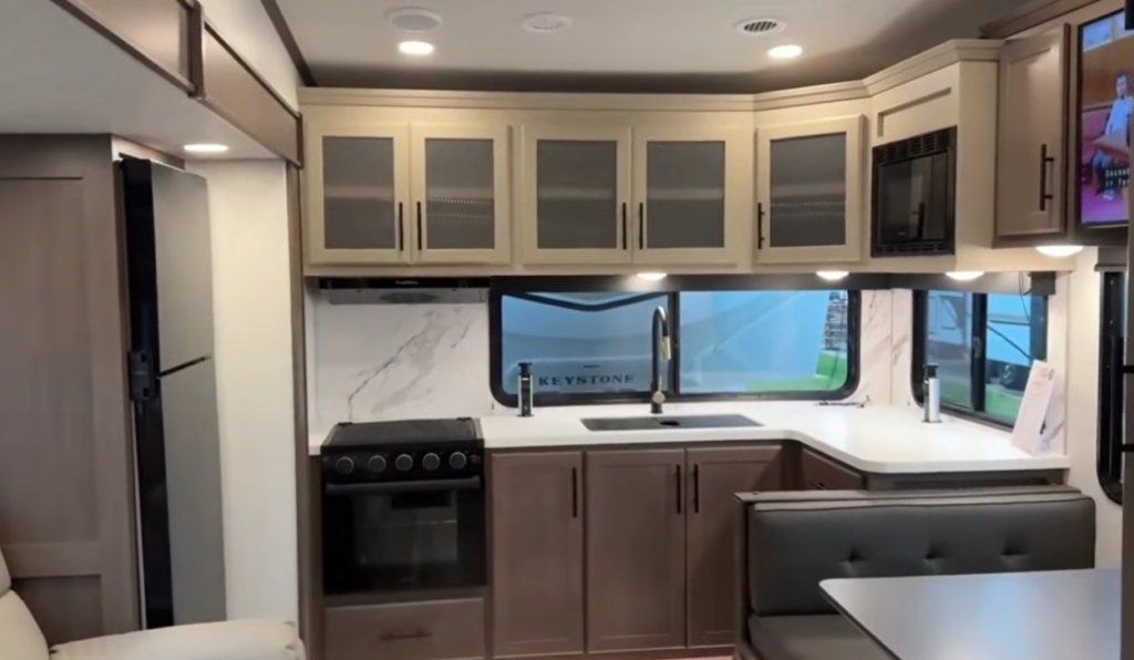 Travel trailer kitchen with L-shaped countertops, large window, overhead cabinets, stove, and adjacent dinette seating