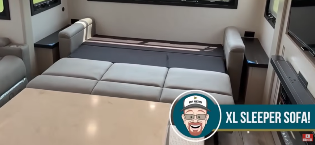 Travel trailer living area with an extended XL sleeper sofa converted into a bed, with text overlay reading “XL sleeper sofa!