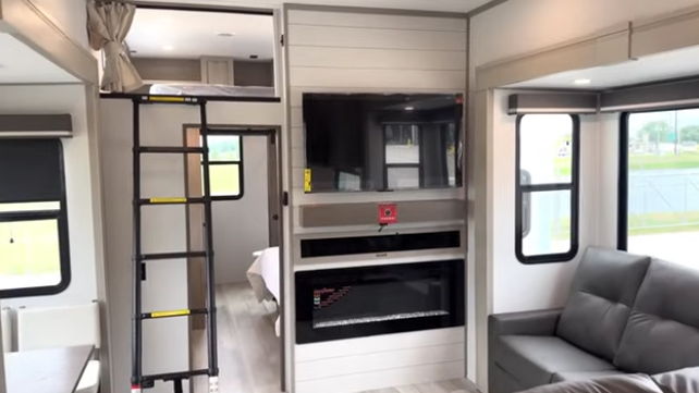 Travel trailer interior featuring an entertainment center with TV and fireplace, ladder access to a loft sleeping area, and adjacent seating