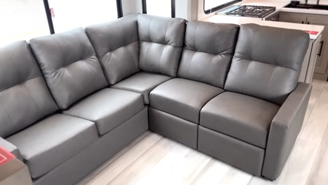 L-shaped sectional sofa inside a travel trailer, featuring gray upholstery and positioned near large windows and the kitchen area.