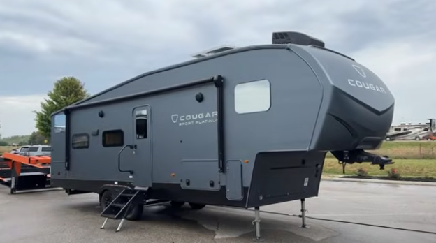 Gray Cougar fifth wheel RV parked on pavement with slide-out extended and front cap visible under cloudy skies.