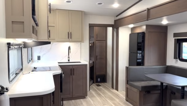 Travel trailer interior with kitchen featuring solid-surface countertops and sink, adjacent dinette seating, and hallway leading to bedroom and bathroom