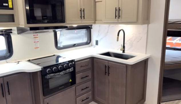 Travel trailer kitchen with L-shaped countertop, sink, stove, microwave, and overhead cabinets, with a bedroom visible through the doorway.