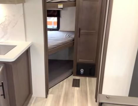 View down a travel trailer hallway into a bedroom with a bed, showing wood cabinetry and vinyl flooring in the foreground.